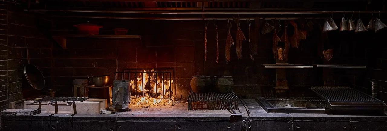 An open fire and grill in a brick kitchen with cured fish hanging above