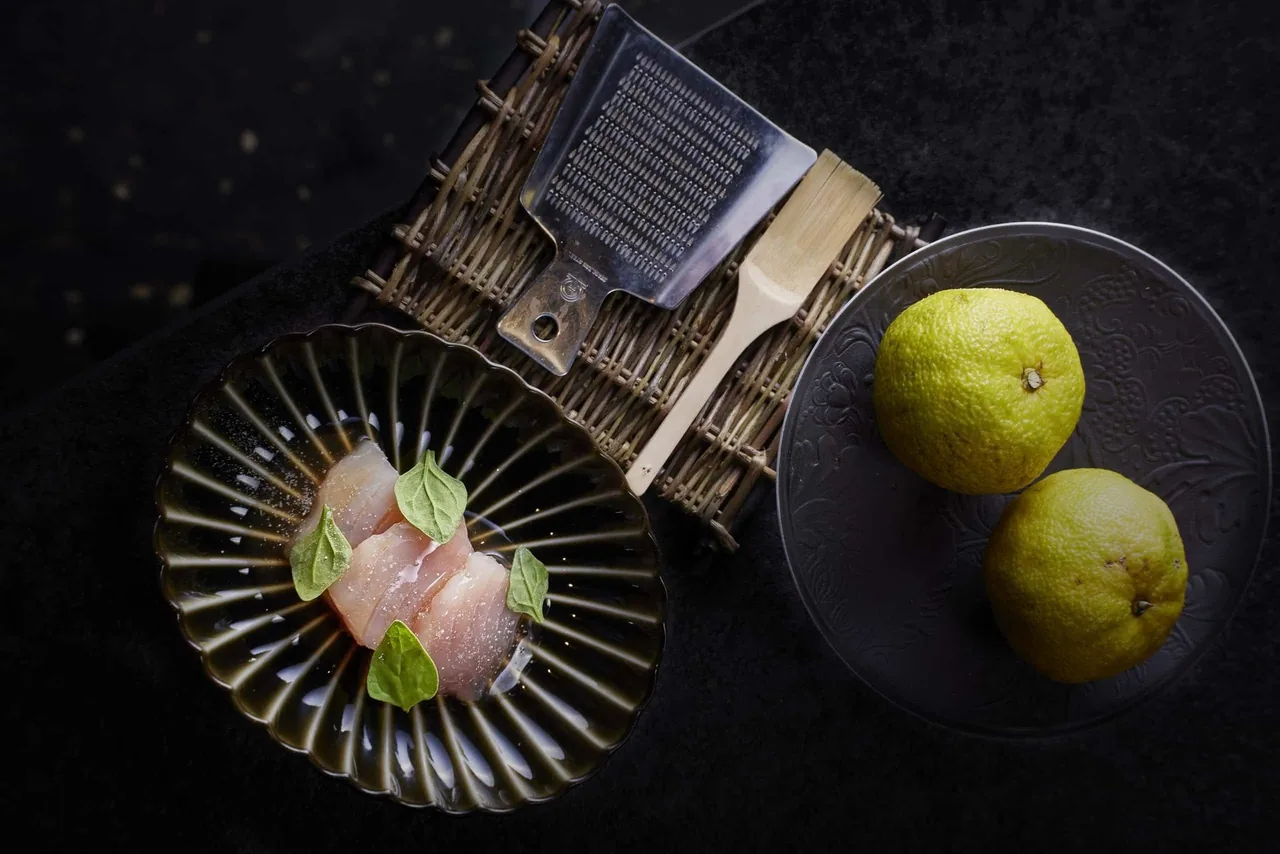 Sliced raw white fish with small green leaves on a fluted dark plate beside two green citrus fruits and a grater