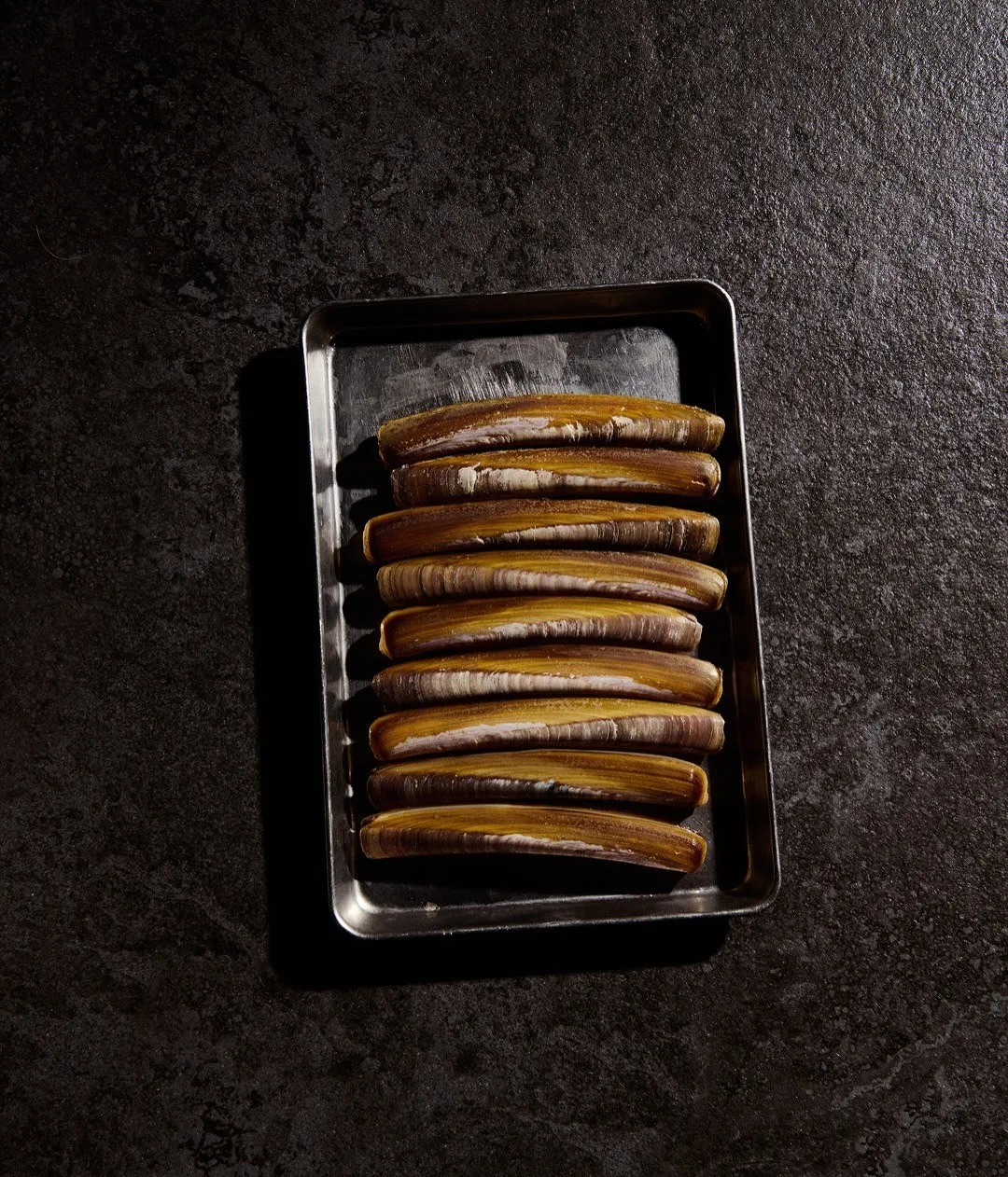 A metal tray of grilled razor clams on a dark stone surface