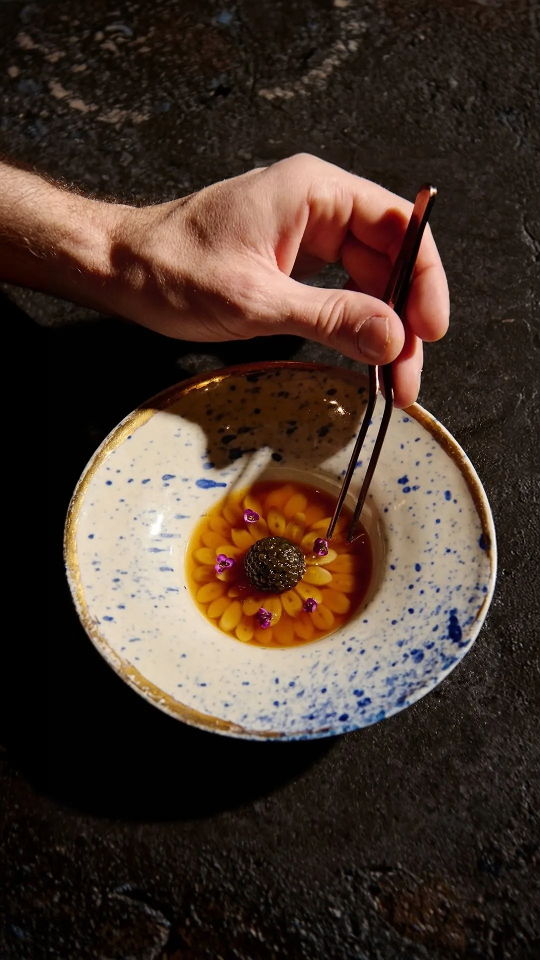 A hand using tweezers over a small speckled bowl holding a glazed dish
