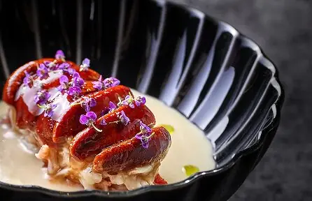 Seared shellfish tails topped with purple edible flowers in sauce, black ridged bowl