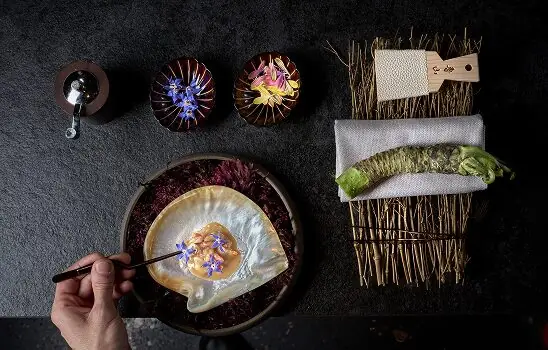 Hand plating a dish with edible flowers on purple leaves, wasabi and small dishes nearby