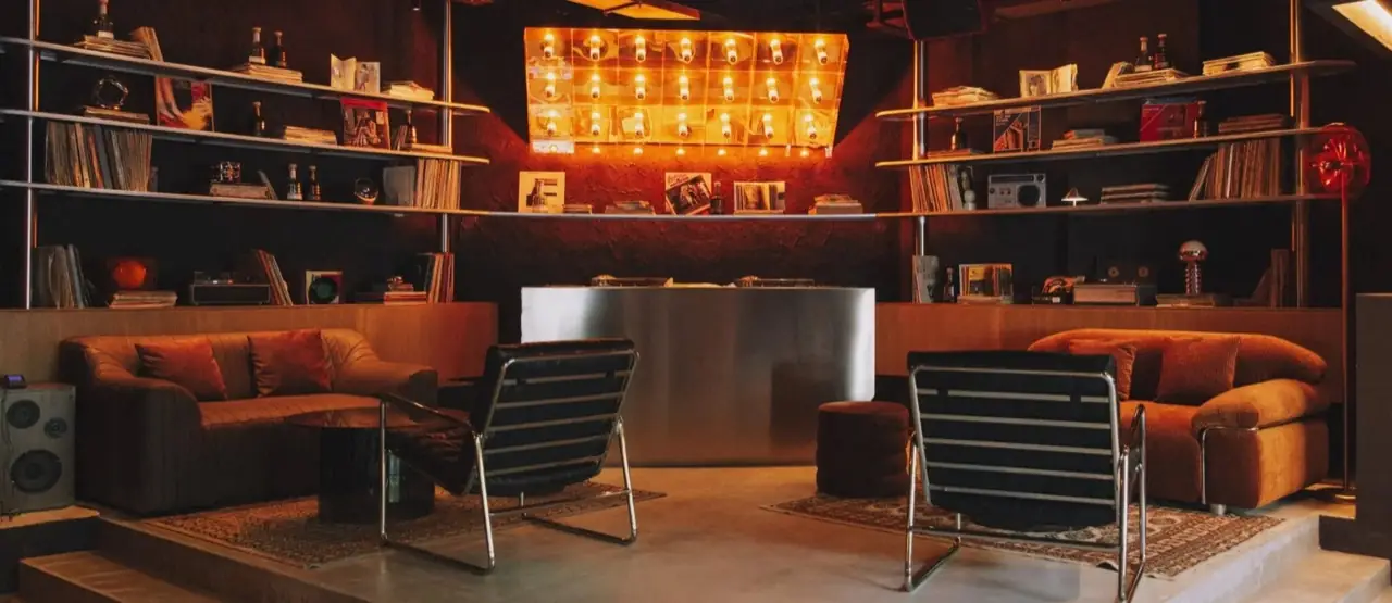 Dimly lit listening-bar lounge with shelves of vinyl records, corduroy sofas and chrome-framed chairs around a steel DJ booth under a backlit panel