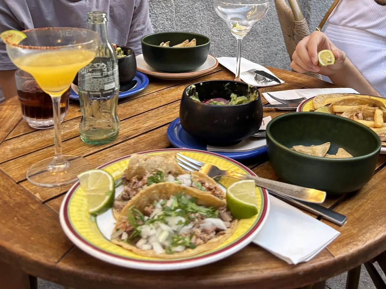A plate of tacos with limes on a wooden table beside a margarita and bowls of side dishes