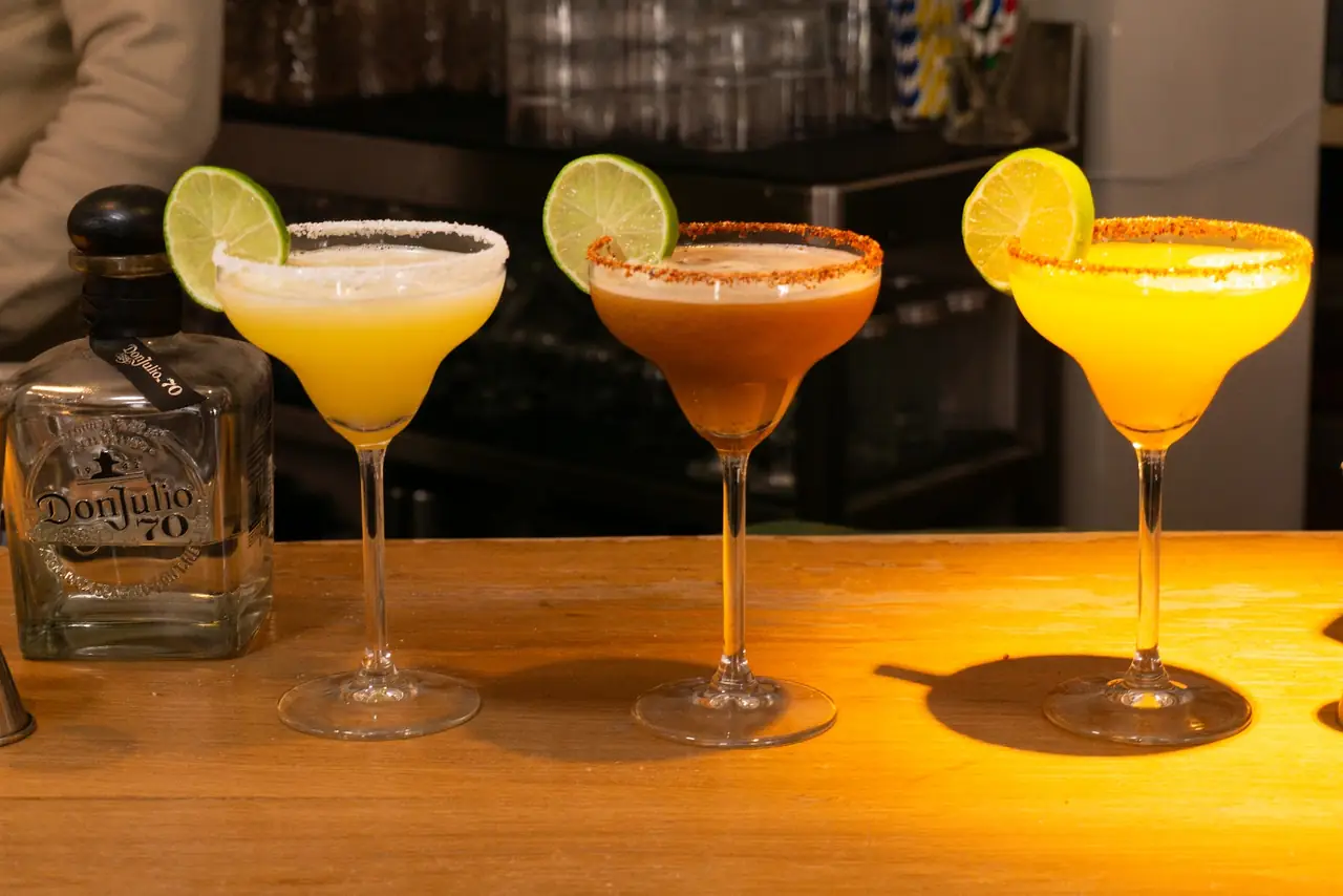 Three margaritas in different flavors with chili-salt and salt rims, garnished with lime, on the bar