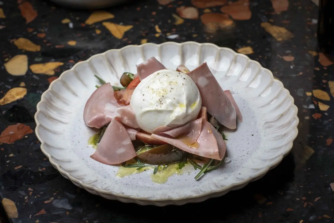 Burrata with sliced ham and olive oil in a scalloped white bowl.
