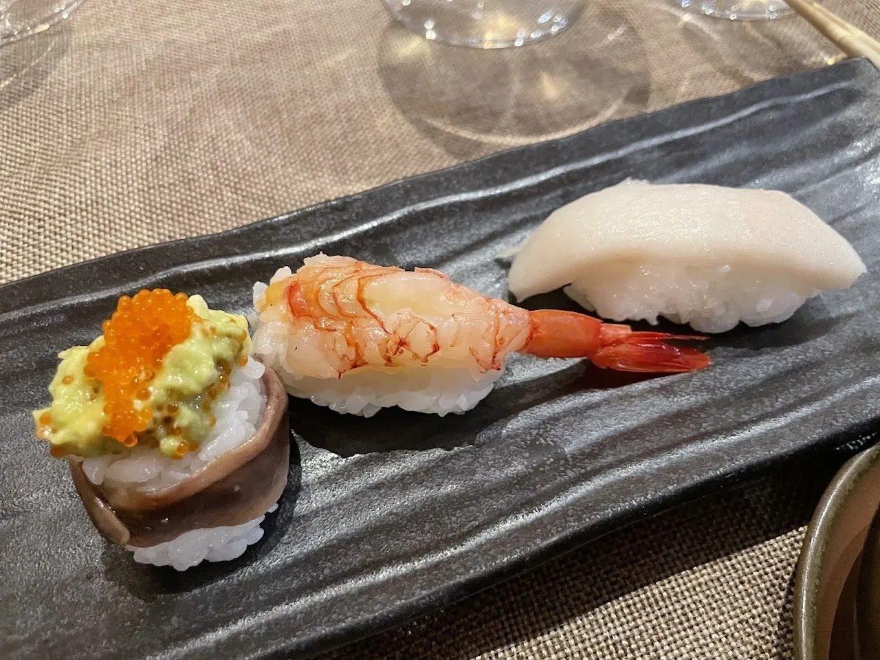 Three pieces of nigiri, one topped with a prawn, on a dark grey plate