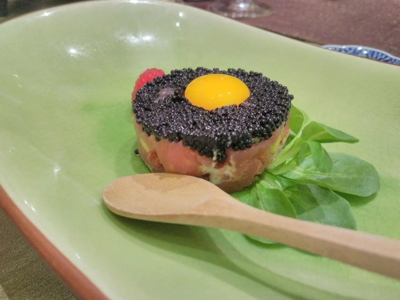A round tartare topped with an egg yolk and black granules on a green plate with a wooden spoon