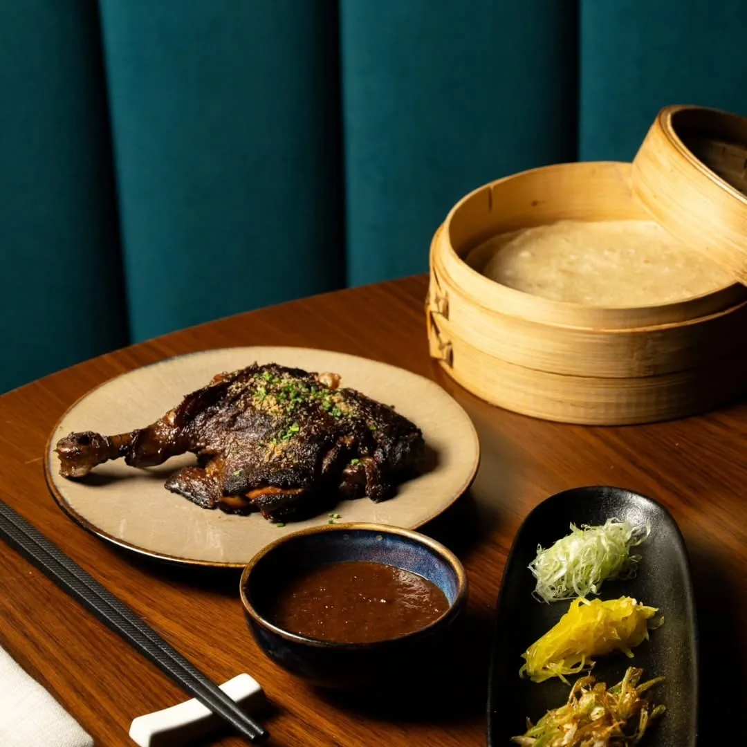 A roast duck leg on a plate beside a bamboo steamer and a small bowl of sauce