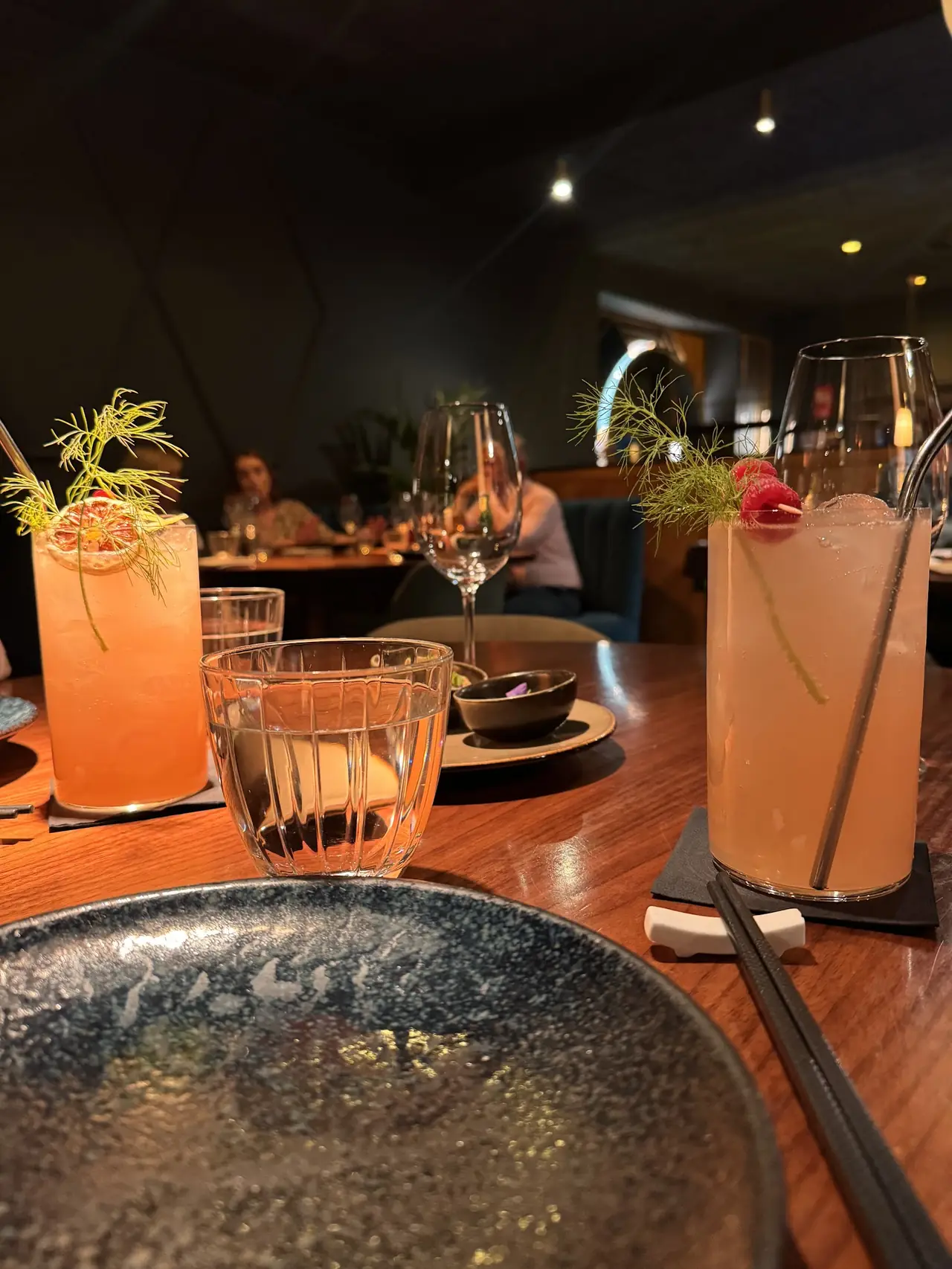Two garnished orange cocktails with fennel fronds and raspberries on a wooden table