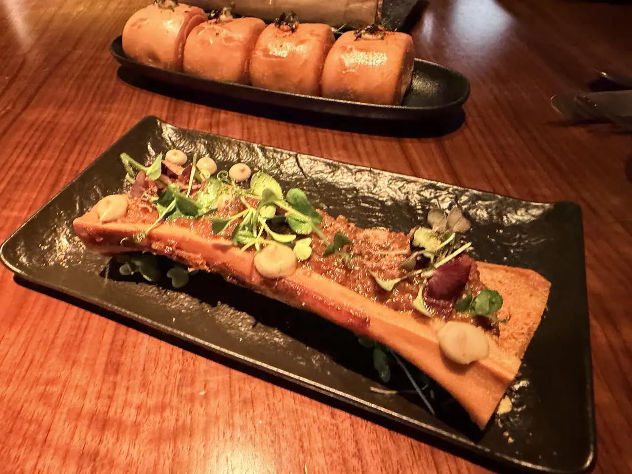 Bone marrow topped with herbs on a black rectangular plate, with buns on a tray behind