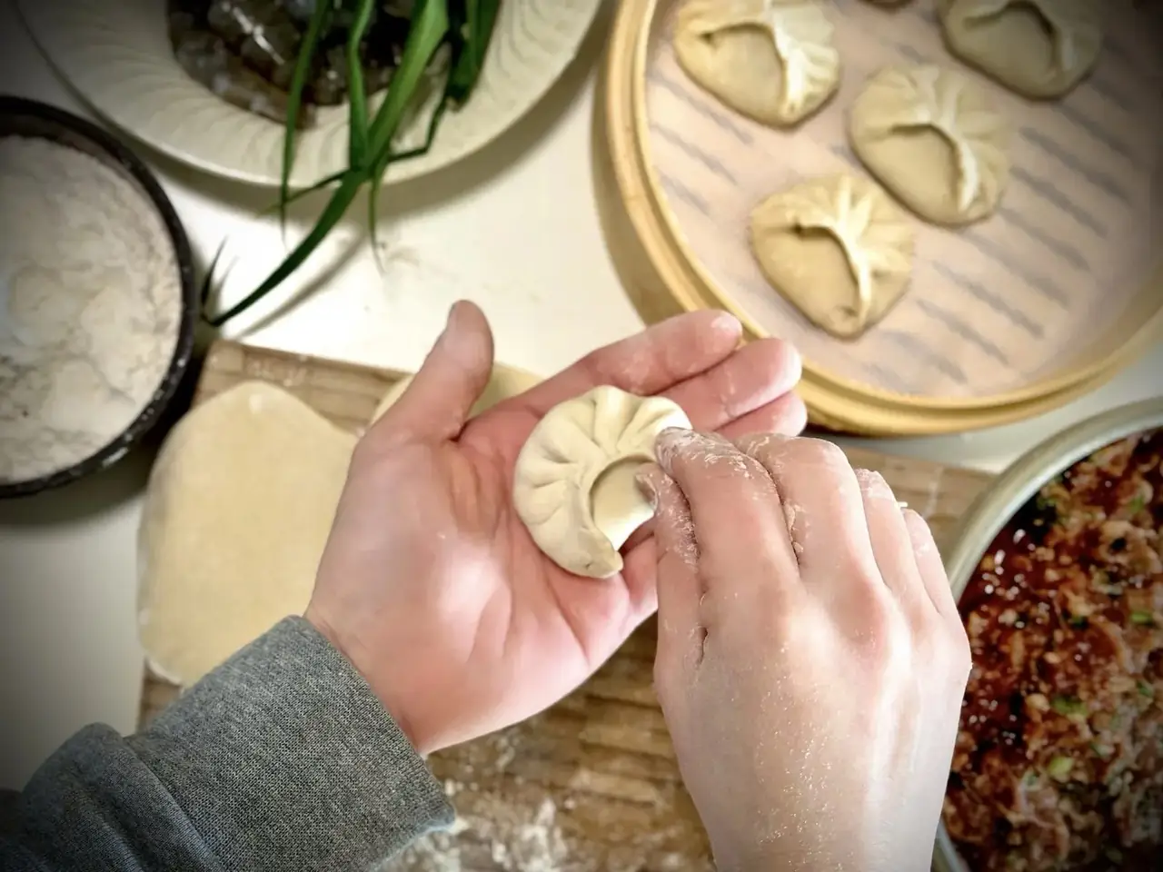 Hands pleating a crescent dumpling by hand, with a bamboo tray of formed dumplings, a bowl of flour, spiced meat filling and greens on a floured board