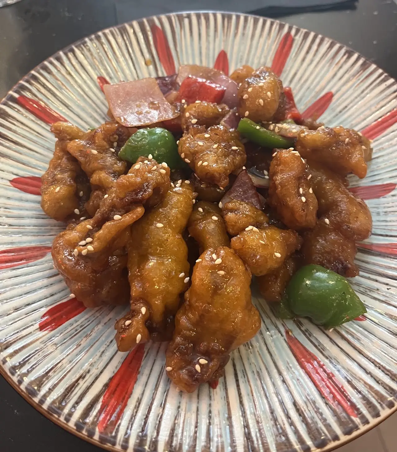 Crispy glazed chicken pieces tossed with red and green peppers, sprinkled with sesame seeds, on a patterned plate.