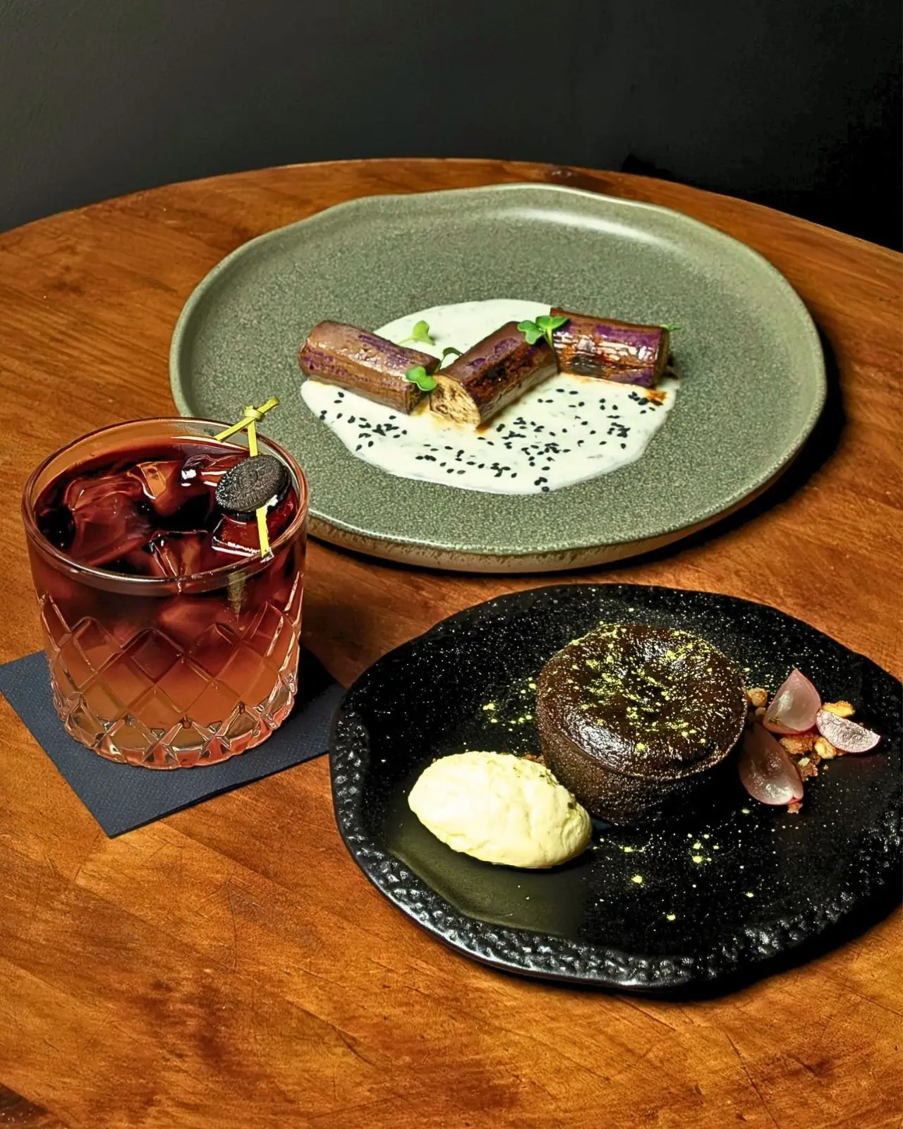 Wooden table with a charred leek dish in white sauce with black sesame on a green plate, a chocolate fondant with cream quenelle and grapes on a black plate, and a red iced cocktail in a cut glass