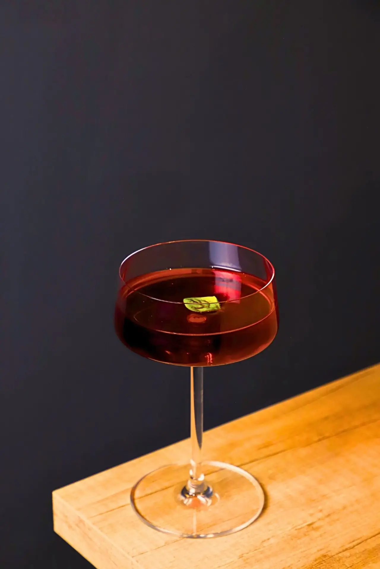 Deep red cocktail in a tall coupe glass garnished with a small green leaf, set on the edge of a wooden ledge against a dark navy wall