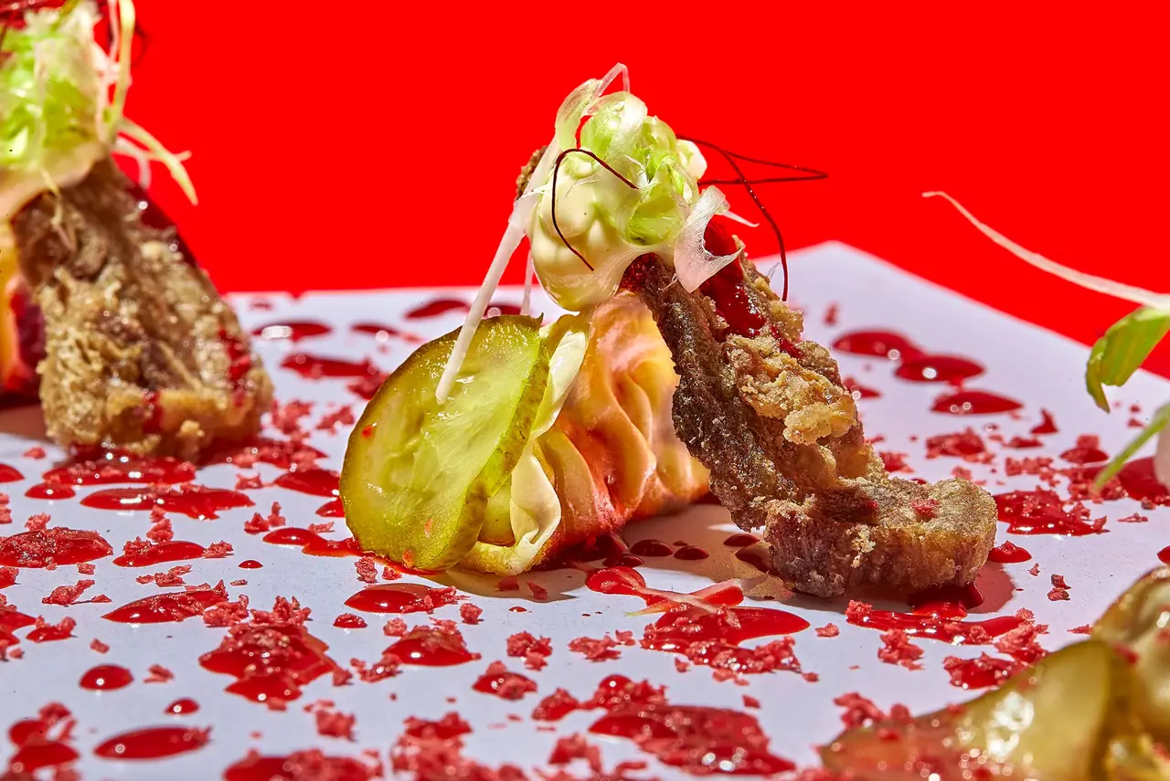Fried cutlet with a pickle slice, folded pale wrapper and shredded greens, scattered with red crumb over a pool of red sauce against a scarlet background.