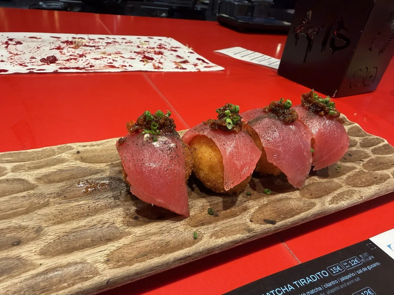 Four pieces of raw tuna draped over round fried croquetas on a carved wooden board on a red counter
