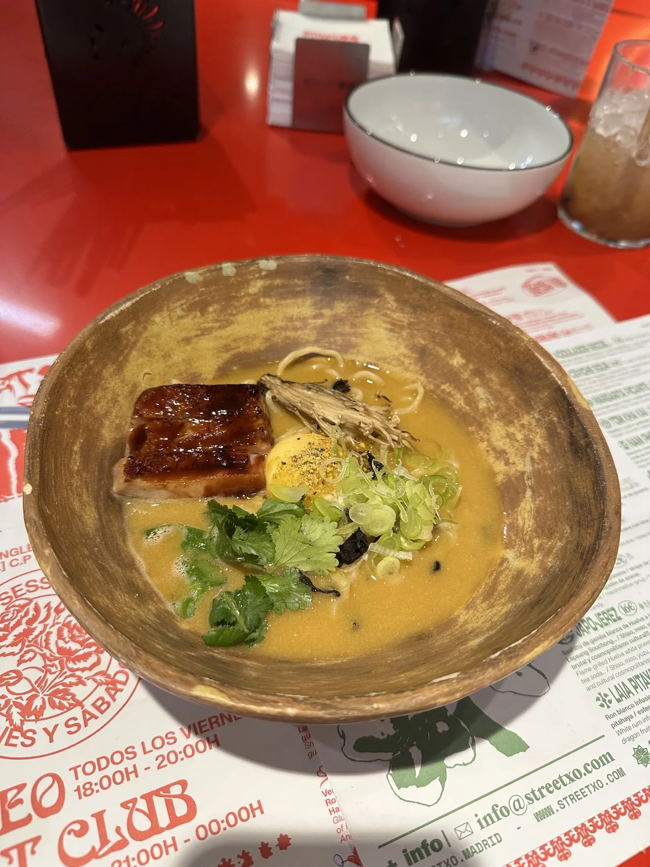 A rustic ceramic bowl of ramen in golden broth with glazed pork belly, an egg yolk, herbs and spring onion