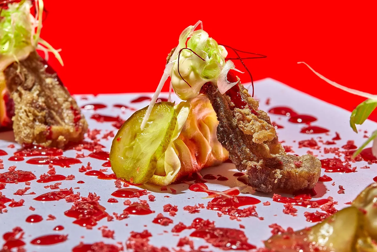 A dumpling and other small bites on white paper with red splatters on a red surface