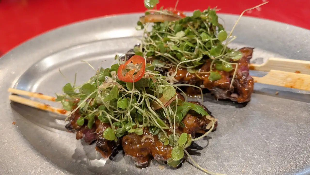 Skewers of grilled glazed meat topped with microgreens and sliced chili on metal plate