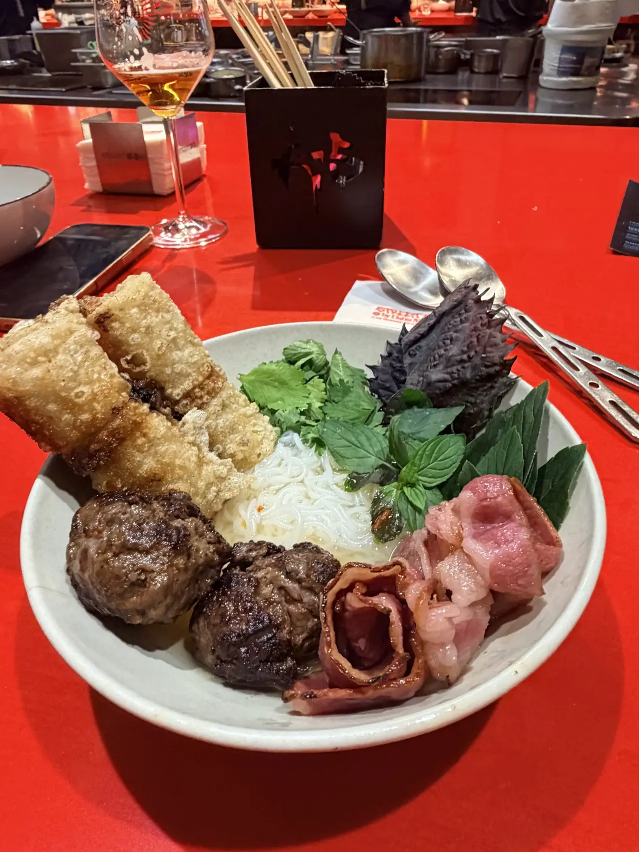 Bowl of noodles with grilled meat, spring rolls, herbs and pork belly, glass of beer behind on red table