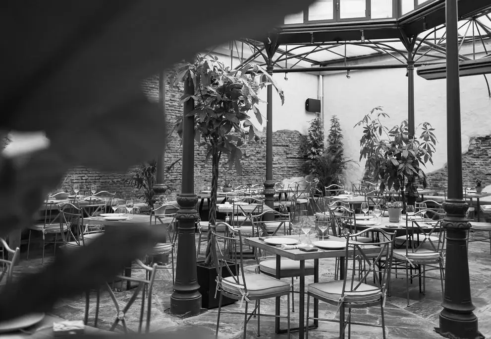 Covered interior courtyard dining room with wrought-iron chairs, marble-top tables and potted trees under an exposed steel-and-glass roof, shot in black and white