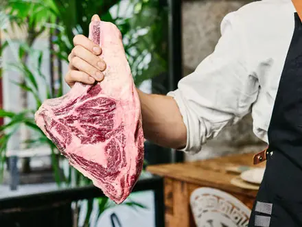 An aproned cook's hand holding up a large marbled raw bone-in beef cut in the dining room