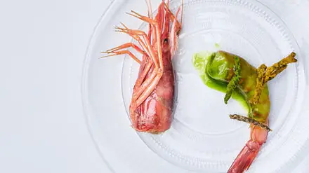 A whole red carabinero prawn plated on a clear glass dish alongside grilled green asparagus in a bright green sauce