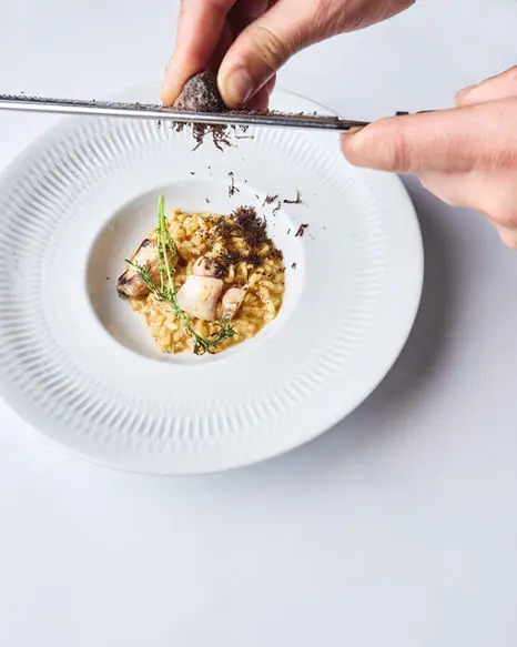 Two hands grating black truffle over a bowl of creamy risotto topped with a thyme sprig