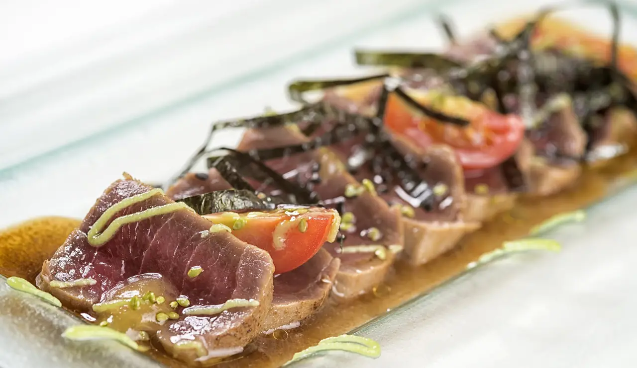 Seared tuna slices with nori strips, cherry tomato, and a green sauce drizzle on a glass plate, at Sucapa