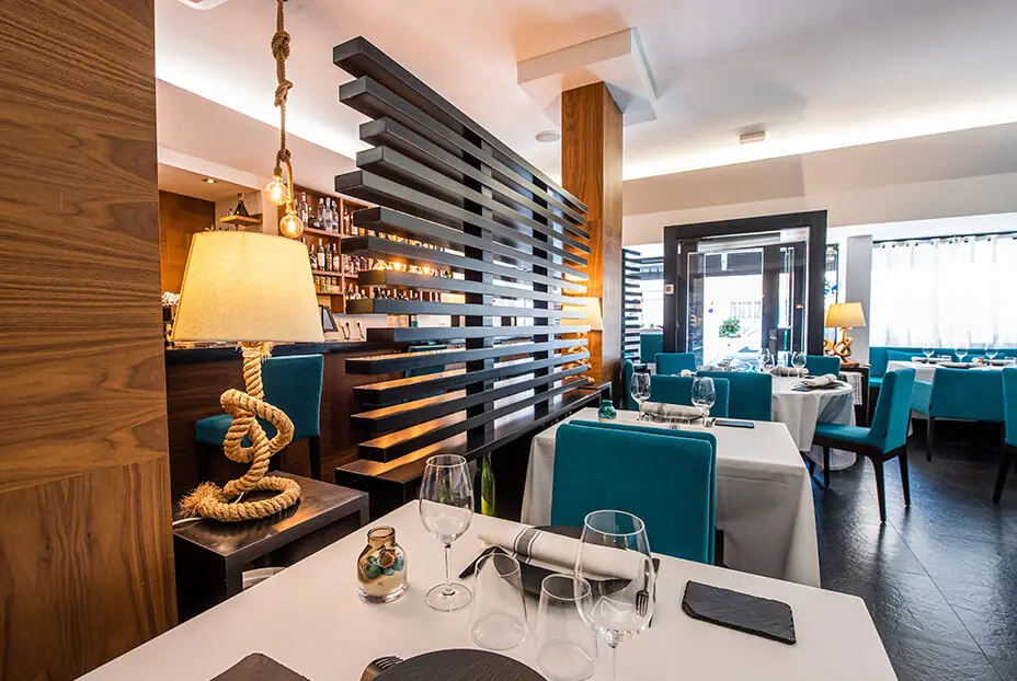A compact dining room with teal velvet chairs and white-clothed tables set with slate mats and wine glasses, walnut panelling, a black horizontal-slat screen dividing the space, a table lamp with a knotted rope base, and the street door and window at the back.