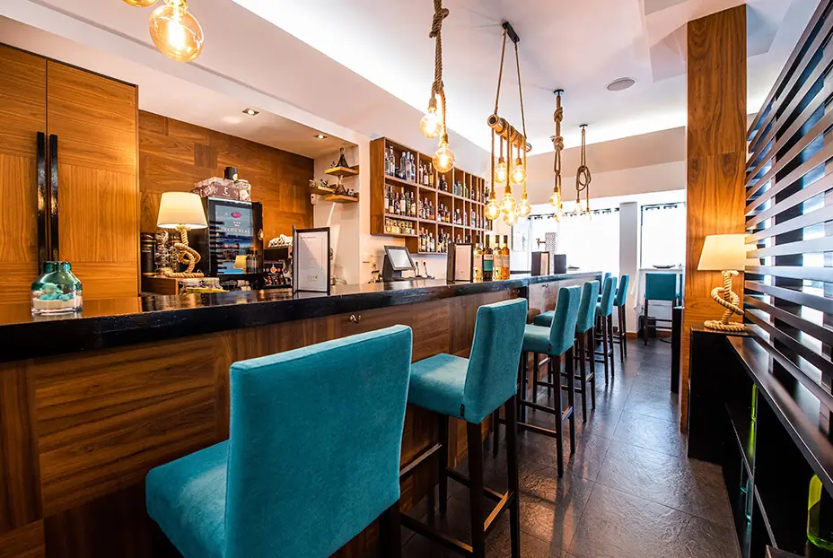 The bar: a long dark stone counter with teal upholstered high stools, walnut panelling, a shelved back bar of spirits, and a cluster of exposed filament bulbs hung on knotted ropes above.