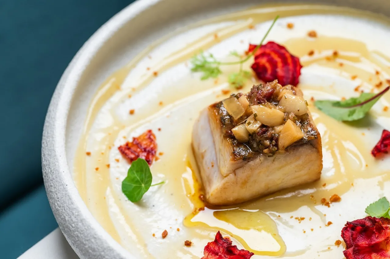 A piece of golden fish topped with nuts on a white plate with red petals and sauce