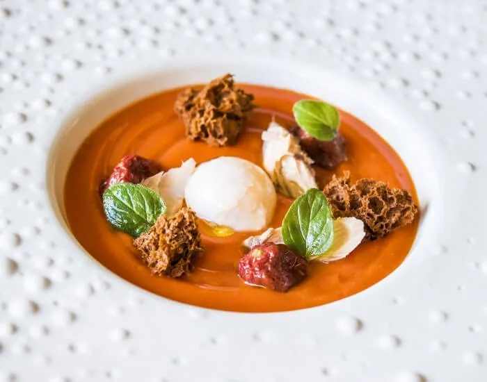 A white plate with orange purée topped with a poached egg, crumbs and basil leaves
