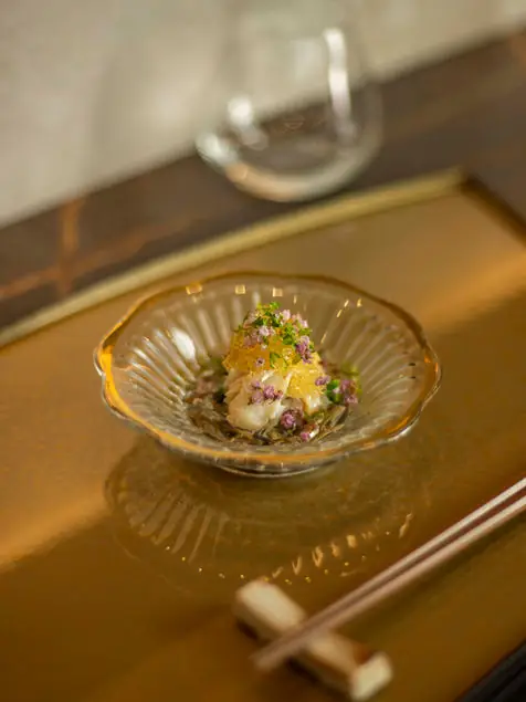 A small Japanese appetizer topped with citrus jelly and purple flowers in a fluted glass bowl on a gold tray with chopsticks