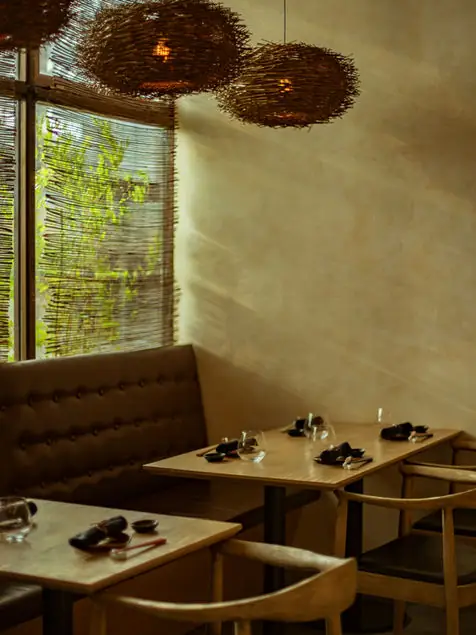 Warmly lit dining room interior with set wooden tables, a leather banquette and woven basket pendant lamps beside a bamboo-screened window