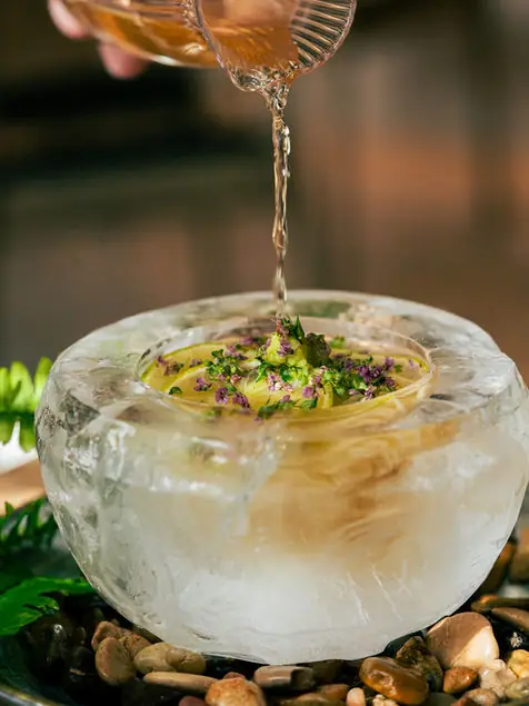 A clear broth being poured from a fluted glass over a dish garnished with herbs and flowers set inside a carved ice bowl on river stones