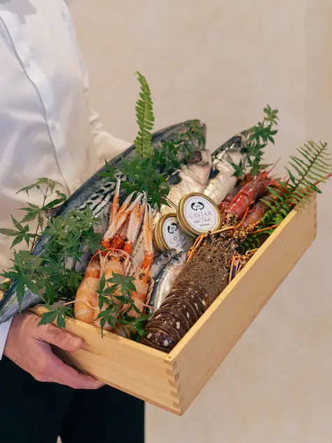 A staff member holding a wooden box displaying whole raw fish, langoustines, spiny lobster and tins of caviar with fern garnish