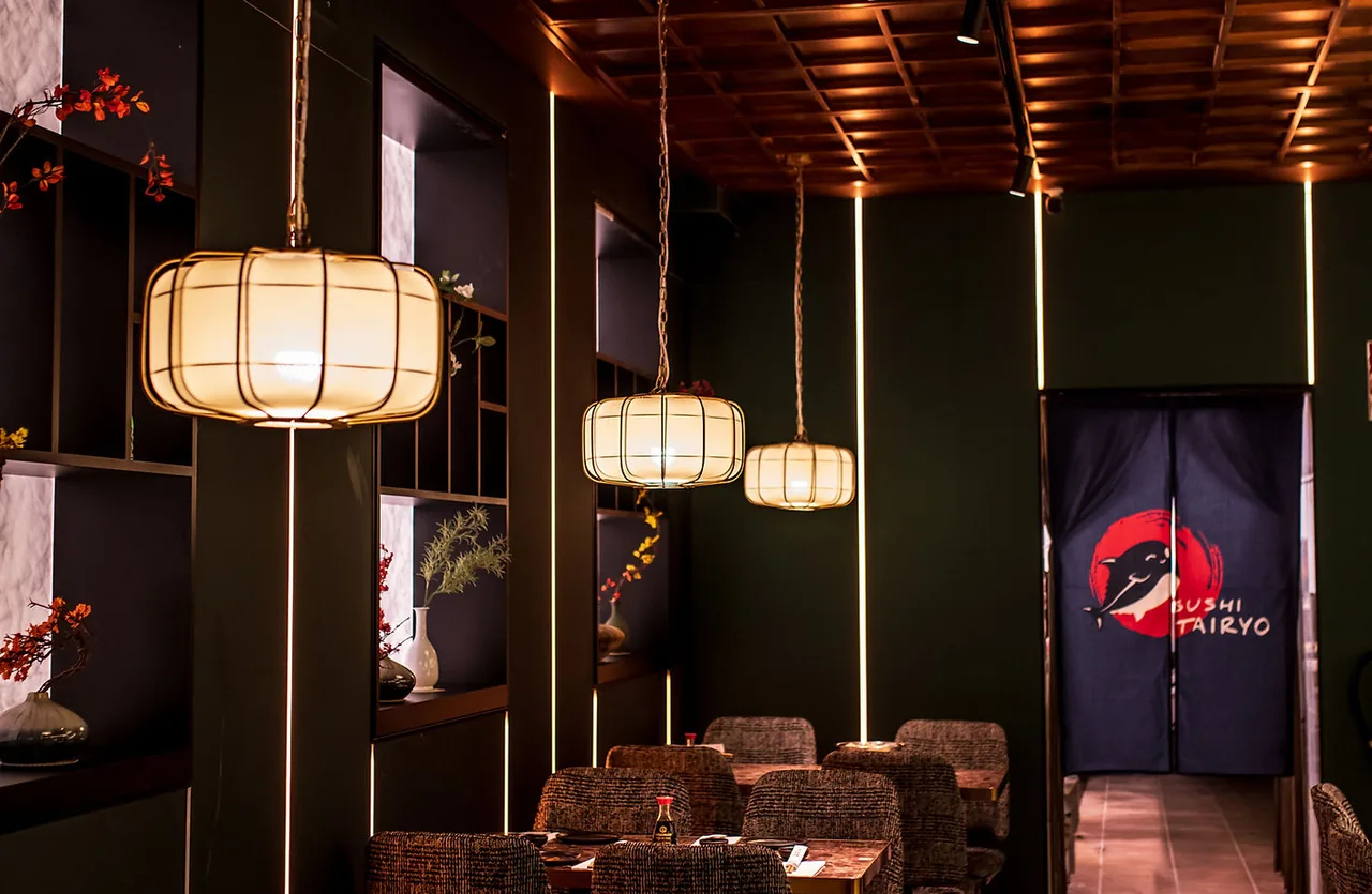 Dark green dining room lit by three cream paper lanterns, woven armchairs at marble-topped tables, and a navy noren curtain printed with the Sushi Tairyo whale-and-red-sun logo over the doorway