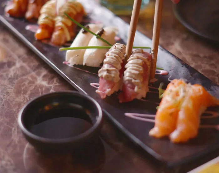 Chopsticks lifting a piece of seared tuna nigiri drizzled with pale sauce from a dark slate board holding salmon and white-fish nigiri, beside a black bowl of soy sauce
