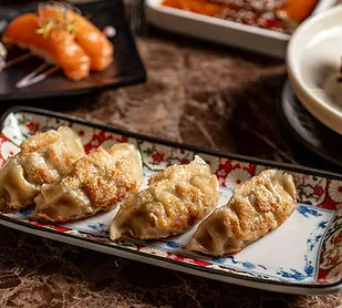 Four crisp-bottomed gyoza lined up on a floral-rimmed rectangular plate on a marble table, with skewers and other dishes blurred behind