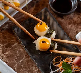 Two nori-wrapped nigiri each topped with a raw quail egg yolk and a dot of black caviar, held between wooden chopsticks over a dark slate board