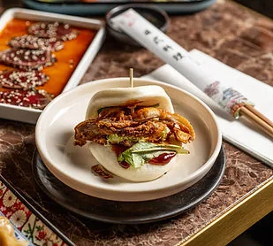 An open steamed bao bun filled with fried chicken, lettuce and red sauce in a small white bowl, next to chopsticks on a printed rest