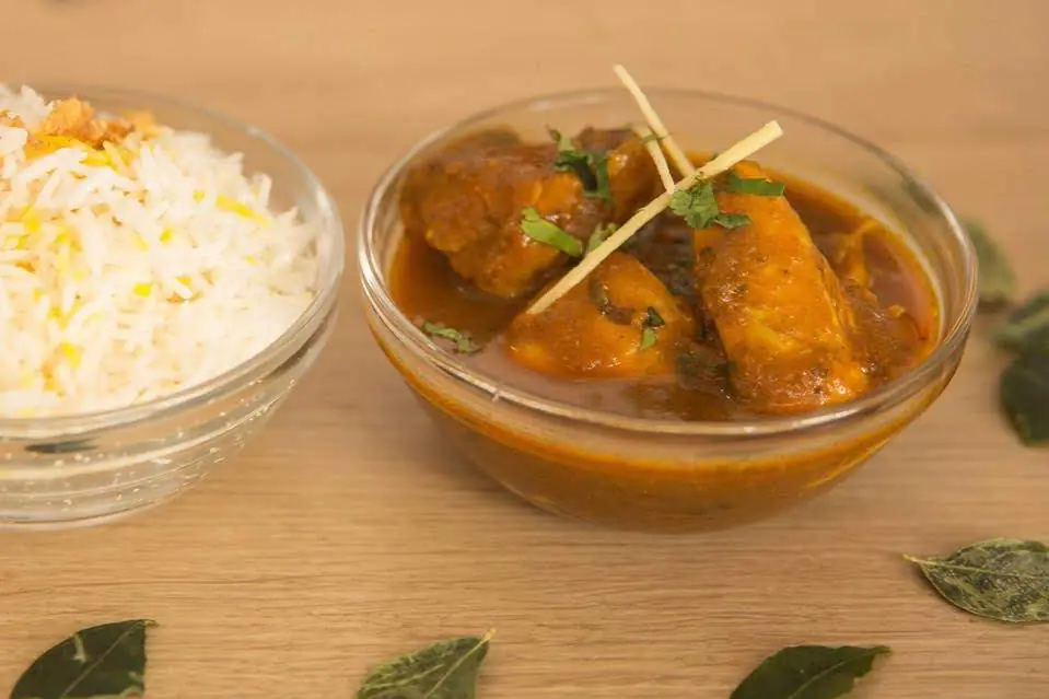 Bowl of chicken curry with cubed meat in a rich tomato-based sauce, garnished with cilantro, served beside a bowl of basmati rice at Swad - The Indian Restaurant