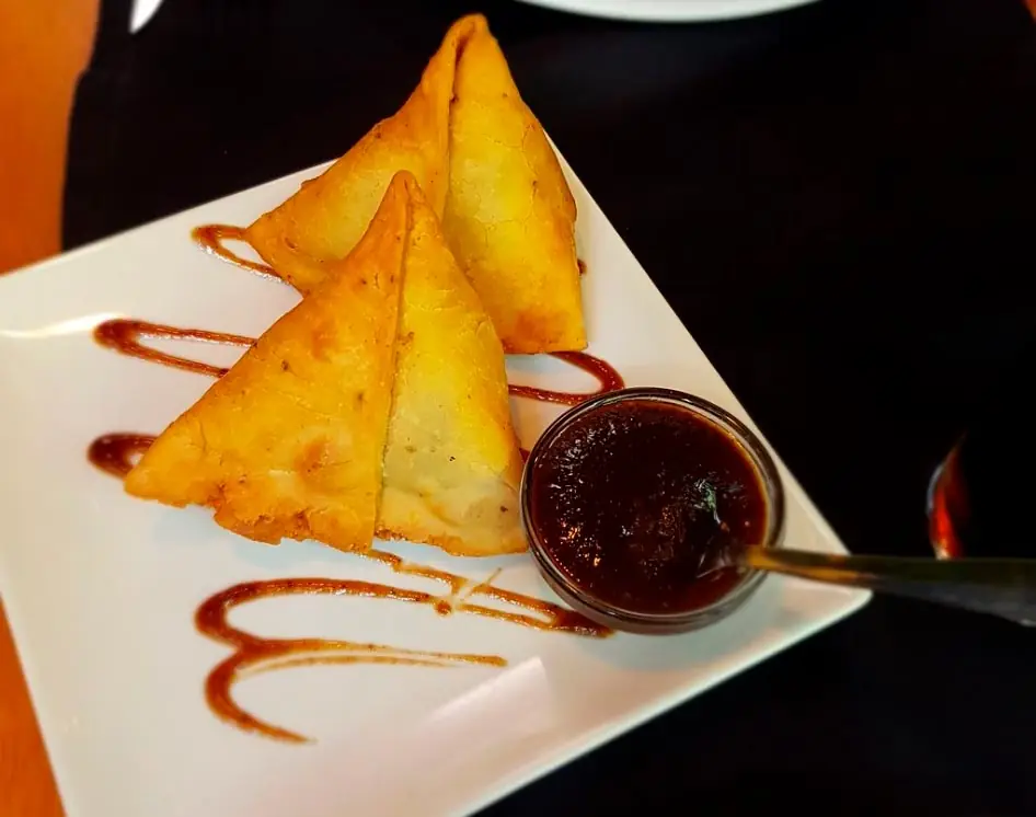 Three fried samosas on a square white plate with tamarind chutney drizzle and a small bowl of dipping sauce at Swad - The Indian Restaurant