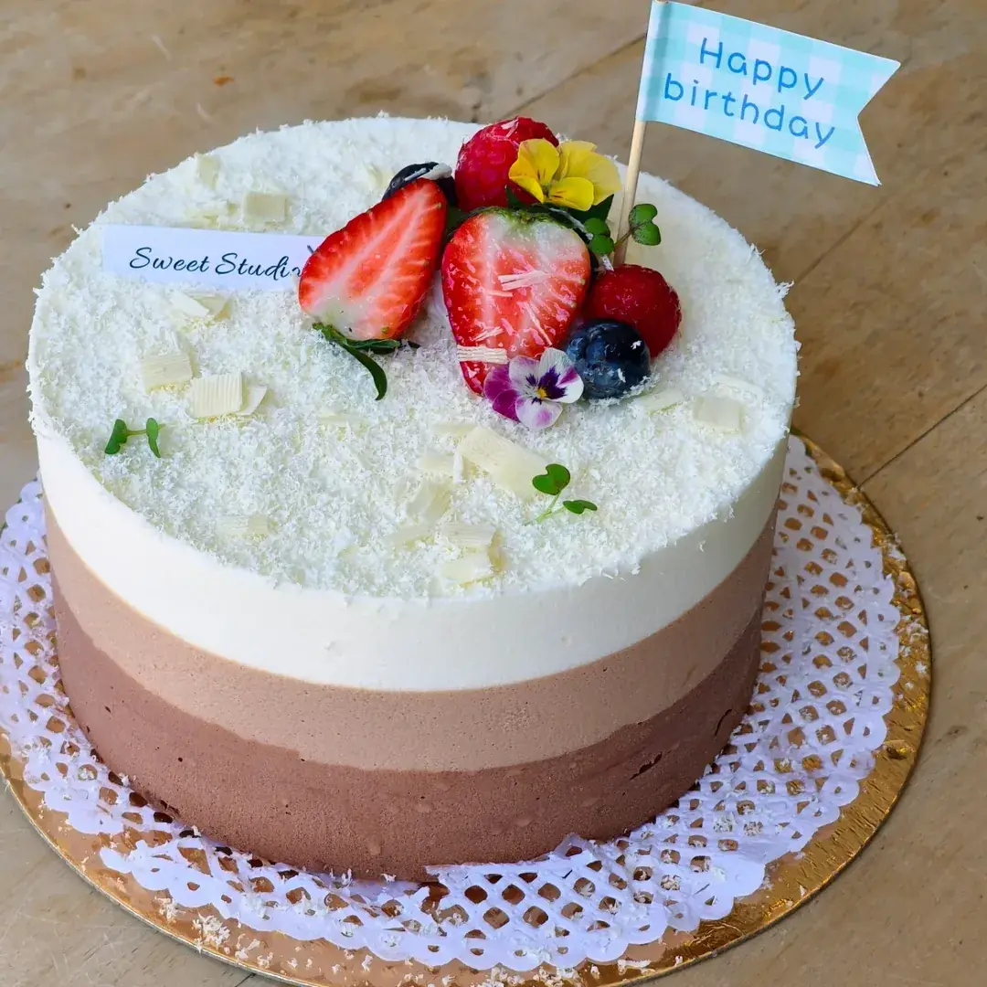 A round cake with white chocolate shavings and fresh berries on top, with a 'Sweet Studio' name card.
