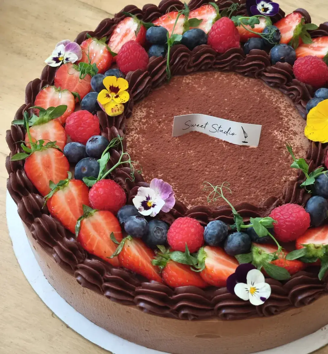 Overhead view of a chocolate cake ringed with berries, edible flowers, and a cocoa-dusted center.
