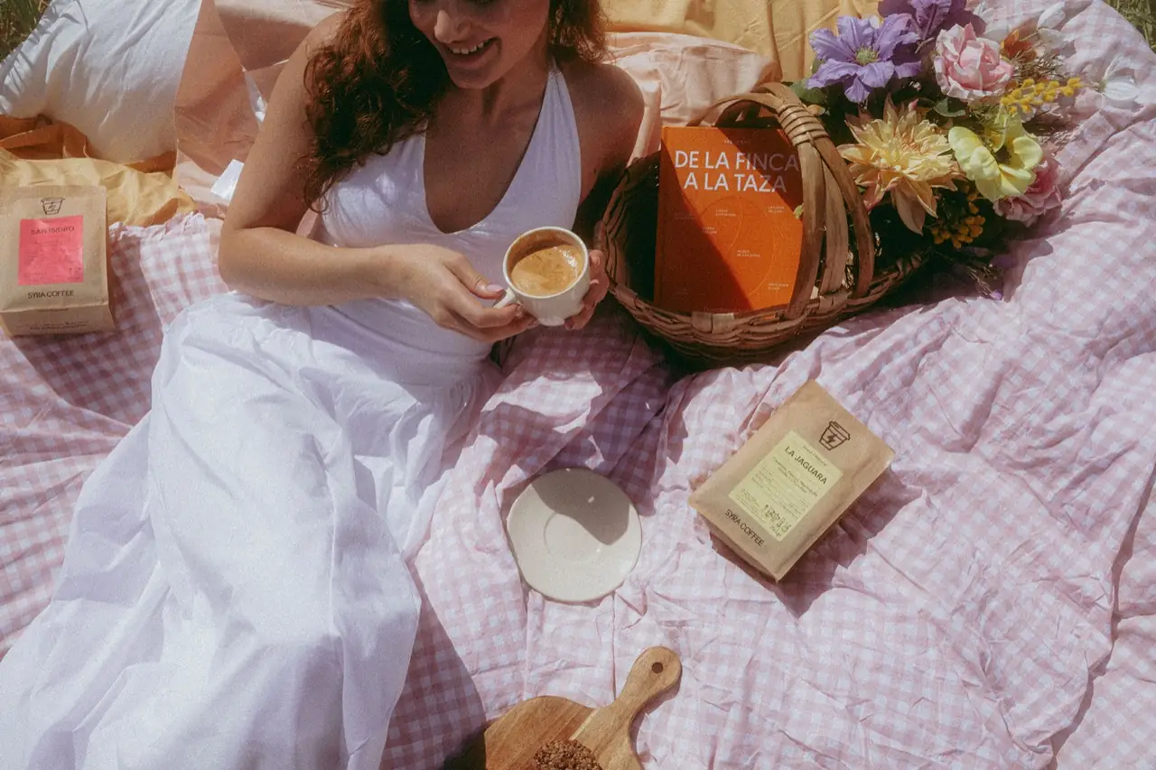 Overhead picnic scene with a served espresso in a white cup and Syra Coffee branded kraft bean bags on a pink gingham blanket