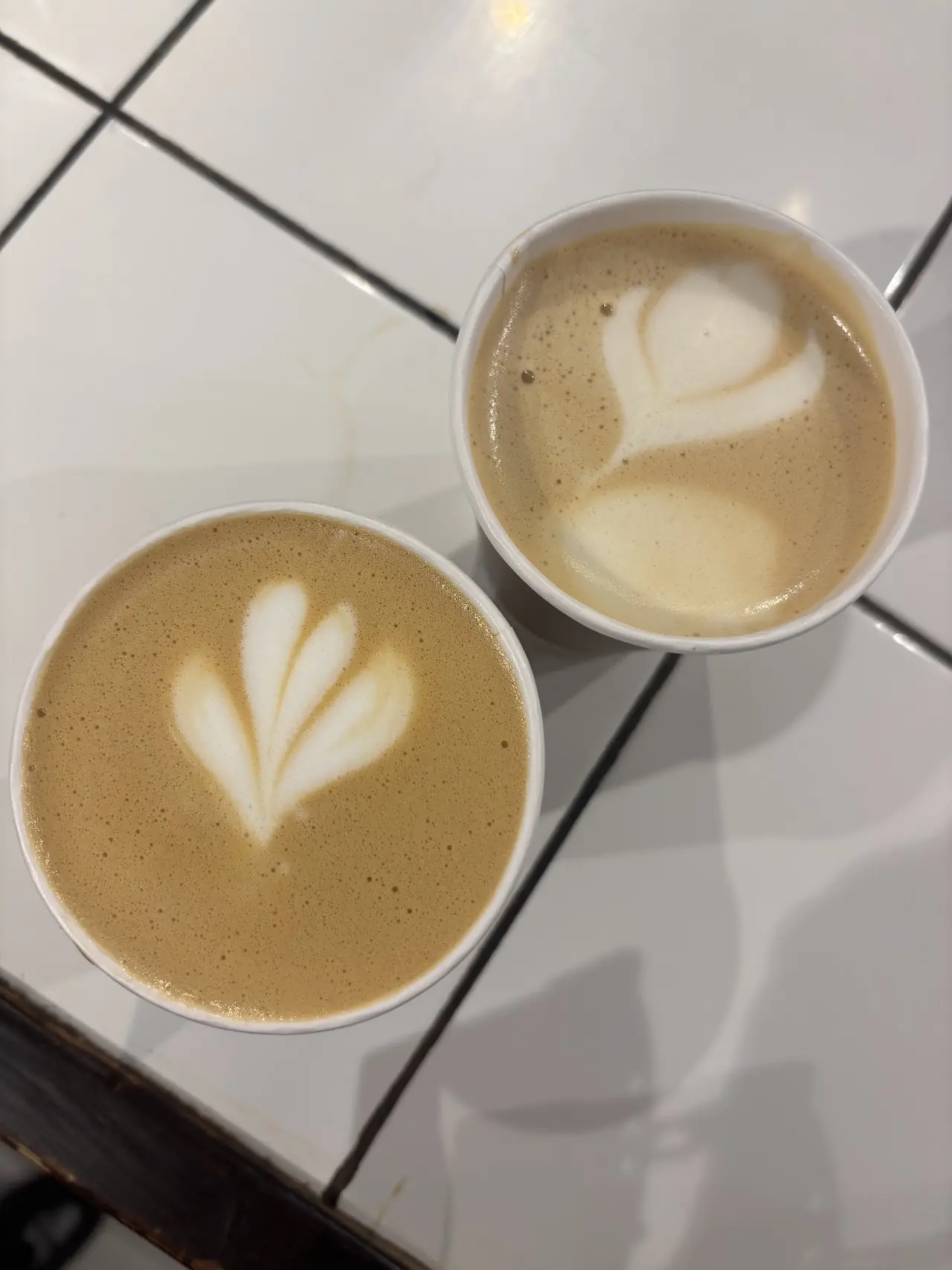 Two lattes with leaf-pattern latte art viewed from above, side by side on a tiled counter.