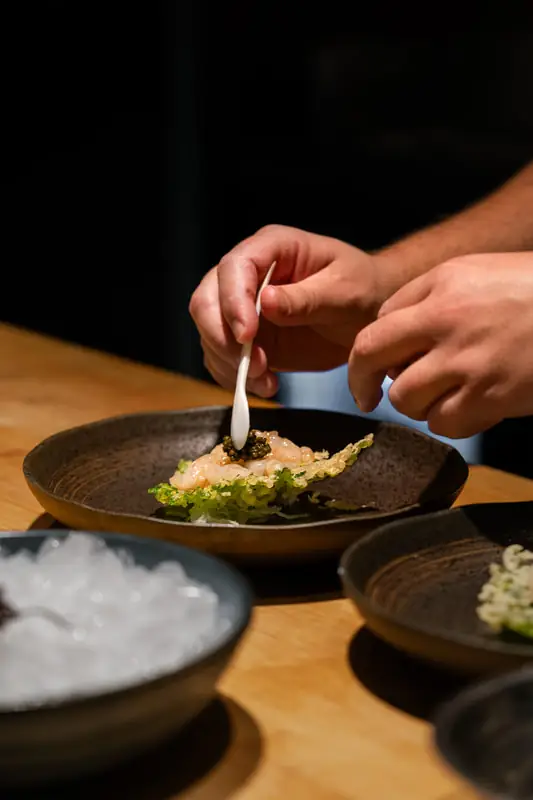 Hands plating a prawn-and-caviar dish with a white spoon in a speckled ceramic bowl beside a bowl of crushed ice